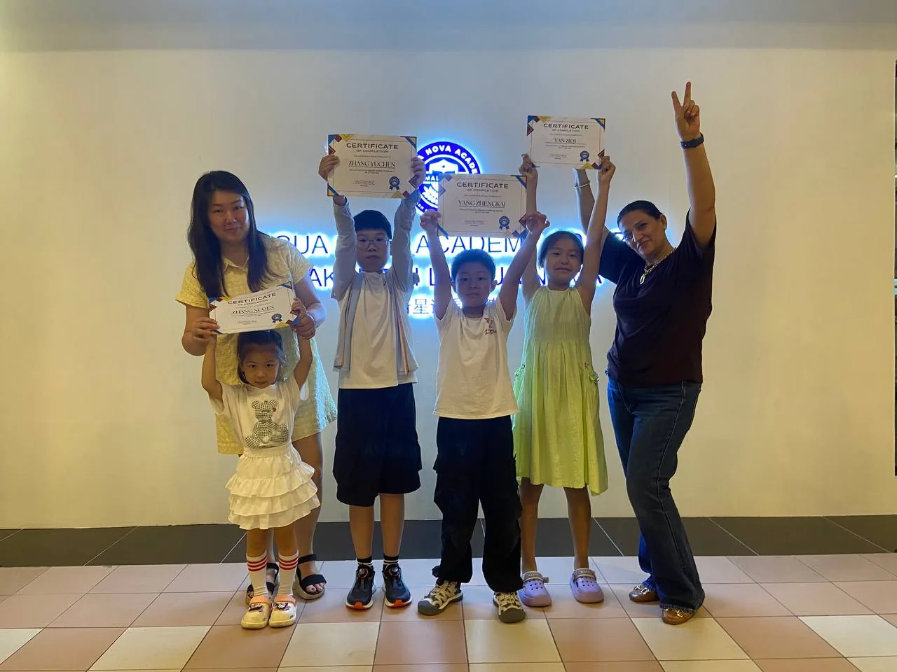 Children holding completion certificates in front of the Lingua Nova Academy sign.