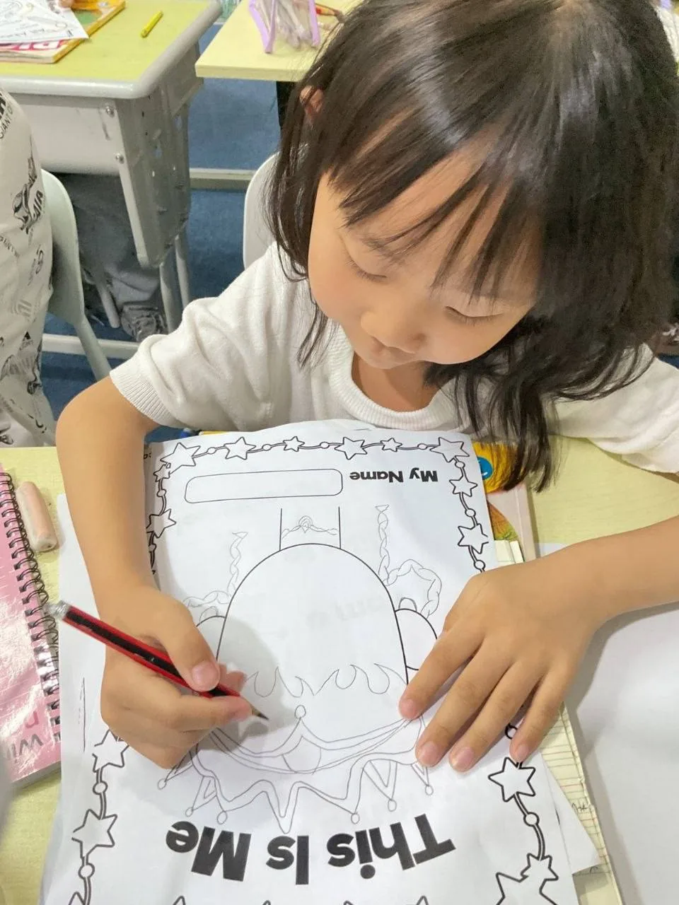 A young learner colouring an English worksheet at her desk.