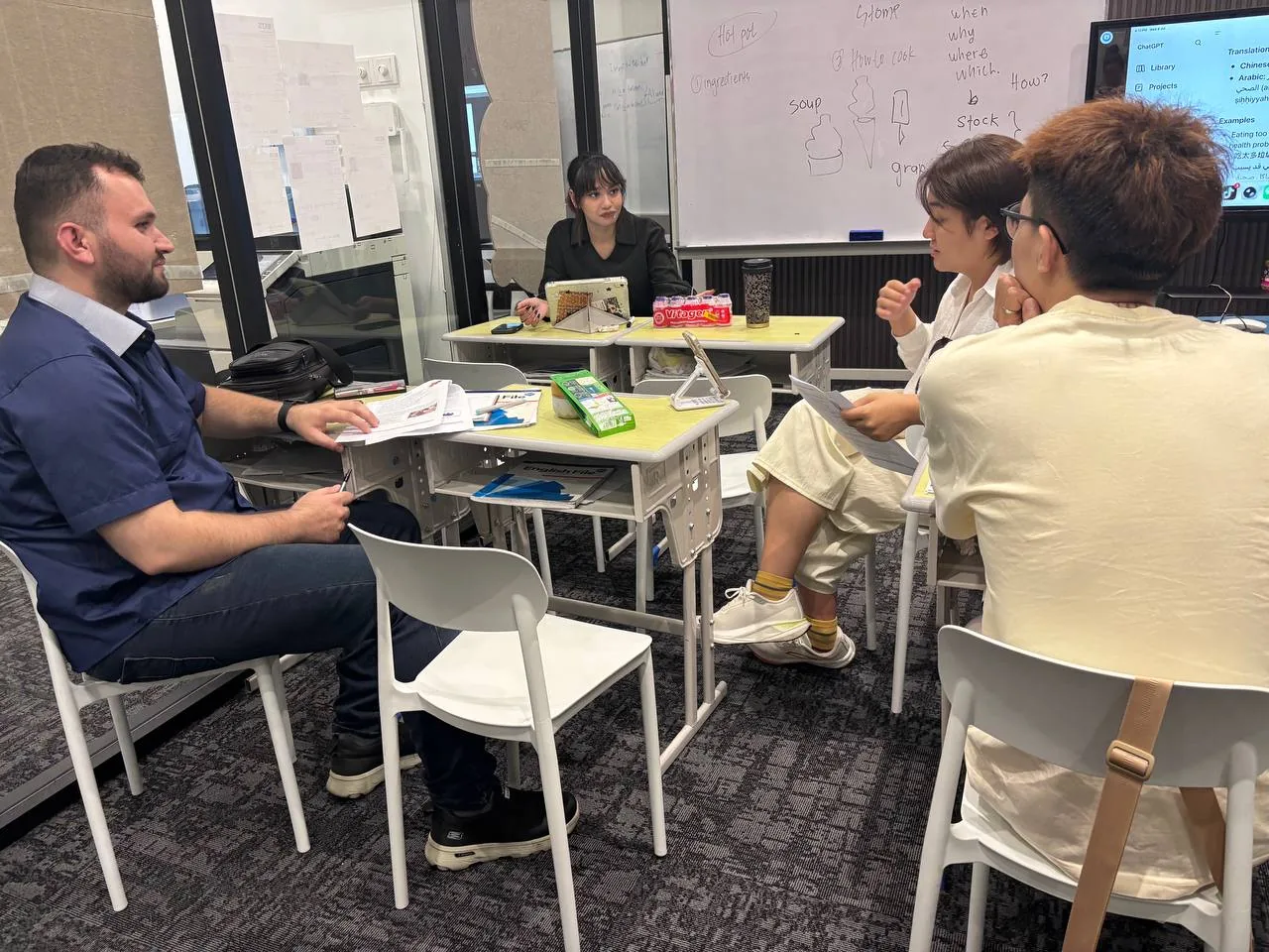 Adult learners and teachers in a small-group English conversation session around a table.