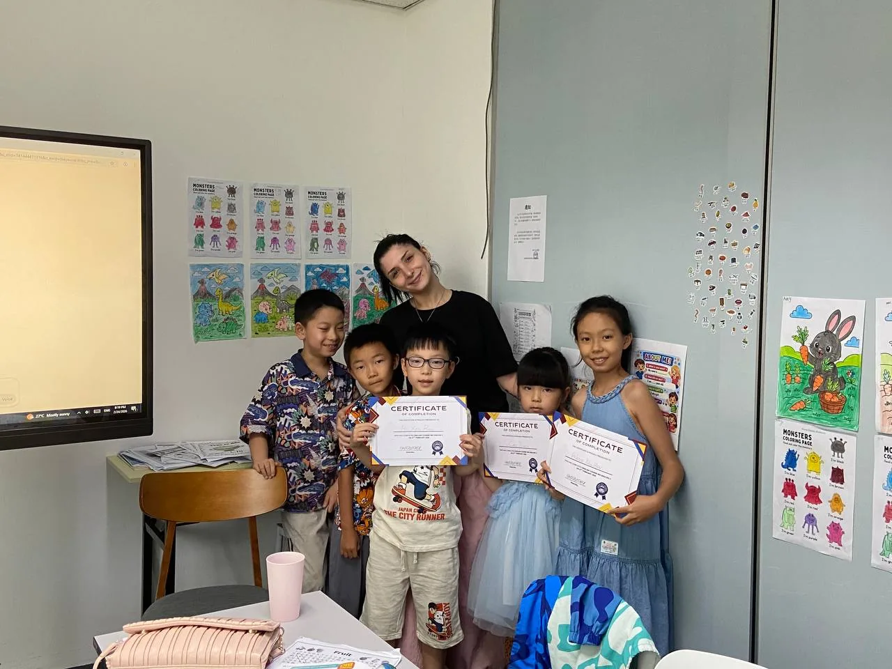 A teacher with a group of young students holding completion certificates in a colourful classroom.